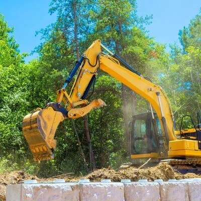 Yellow tracked excavator with extended boom removing soil on commercial construction site surrounded by trees